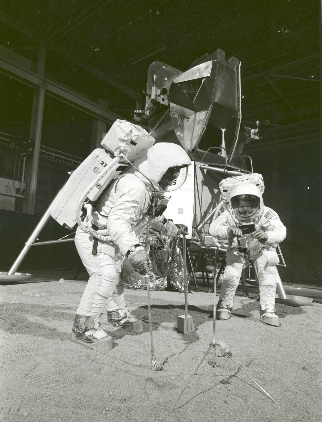 Two members of the Apollo 11 lunar landing mission participate in a simulation of deploying and using lunar tools on the surface of the Moon during a training exercise on April 22, 1969. Astronaut Buzz (Aldrin Jr. on left), lunar module pilot, uses a scoop and tongs to pick up a soil sample. Astronaut Neil A. Armstrong, Apollo 11 commander, holds a bag to receive the sample. In the background is a Lunar Module mockup.